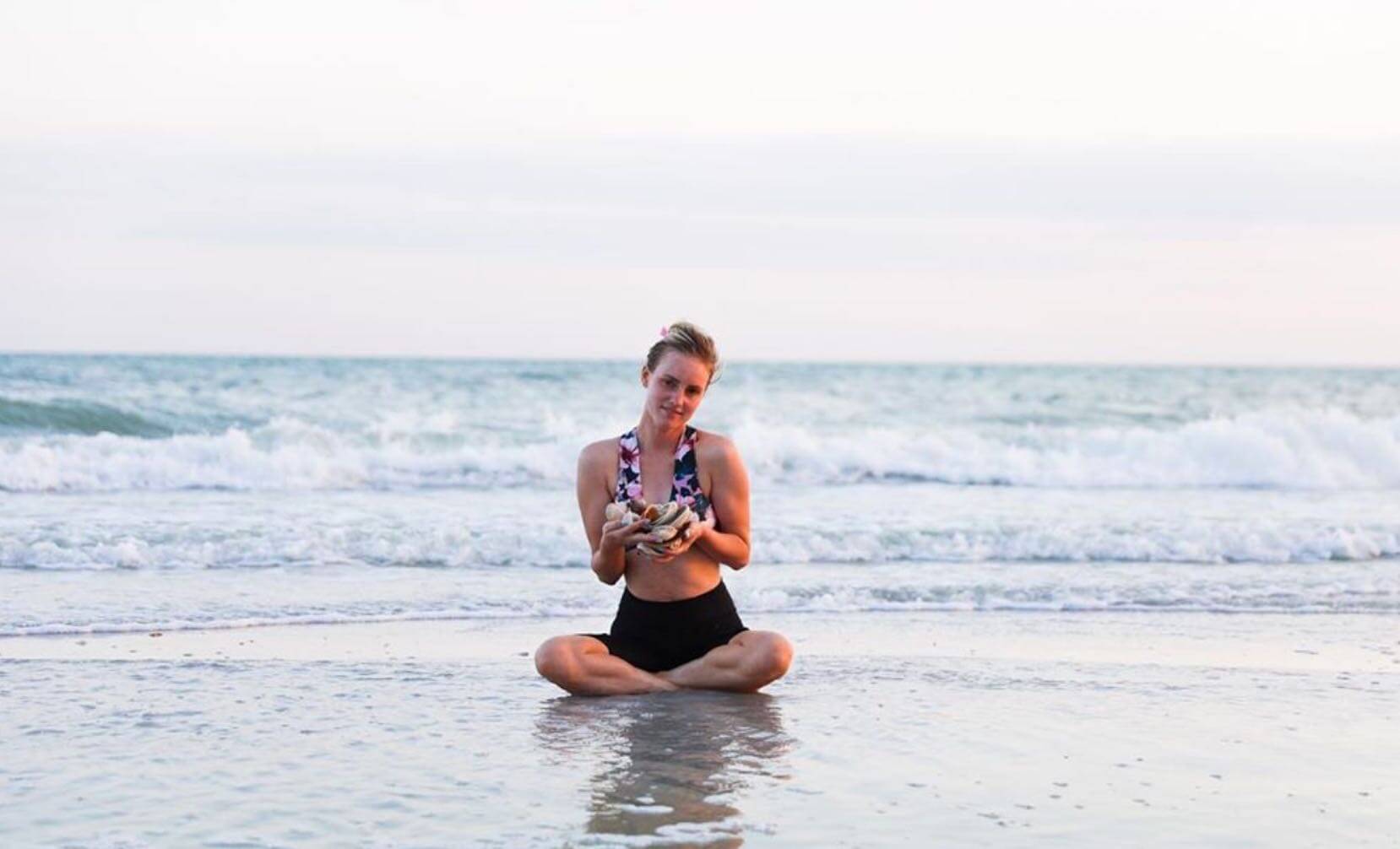 woman sits in shore of beach on sanibel holding shells